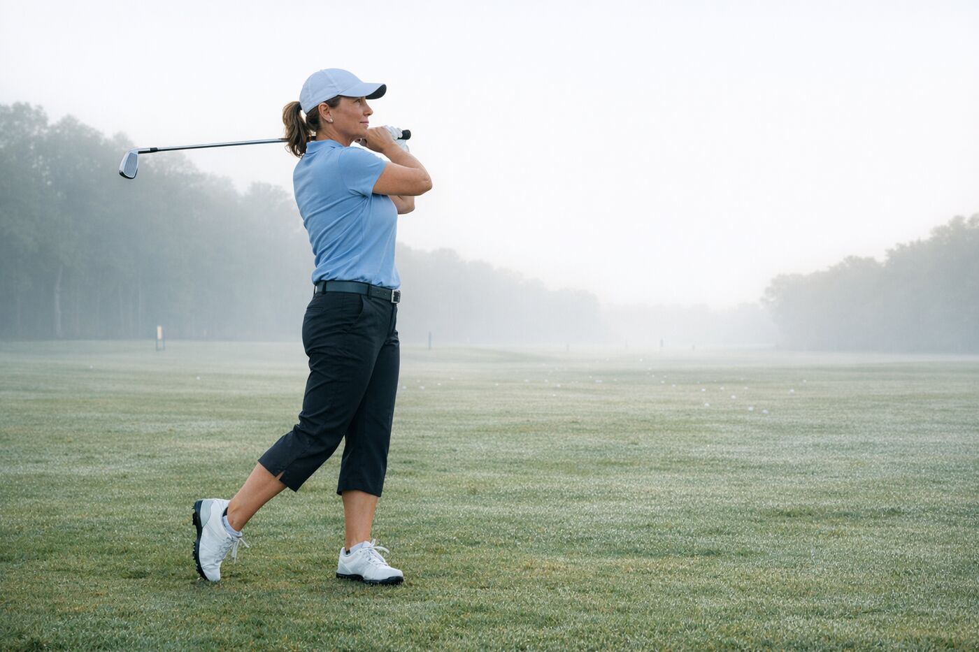 A woman golfer holds a balanced full finish with an iron on a misty driving range in the early morning, the club wrapped behind her neck and her back foot up on its toe.